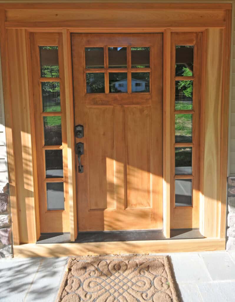 A wooden front door with six small square windows at the top and glass side panels, letting in sunlight. A patterned welcome mat lies on the stone doorstep. The scene is bright and inviting.