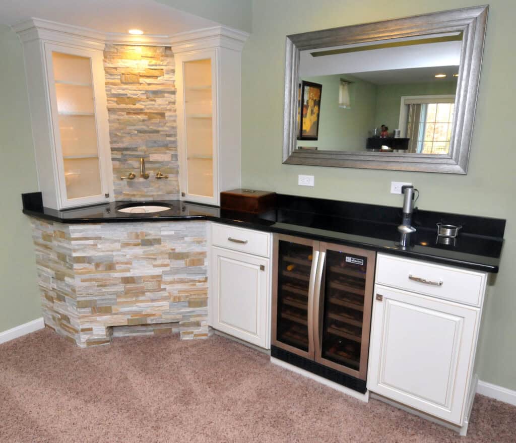 A small home bar area with a sink, stone accent backsplash, black countertop, white cabinets, a wine fridge, and a mirror above. The bar is set against a pale green wall on beige carpet.