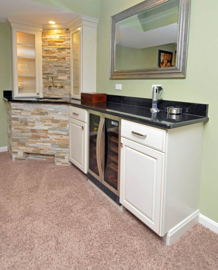 A stylish home bar area with cream cabinets, a black countertop, a built-in wine fridge, a large mirror, and a stone accent wall with a sink, all set on tan carpet.