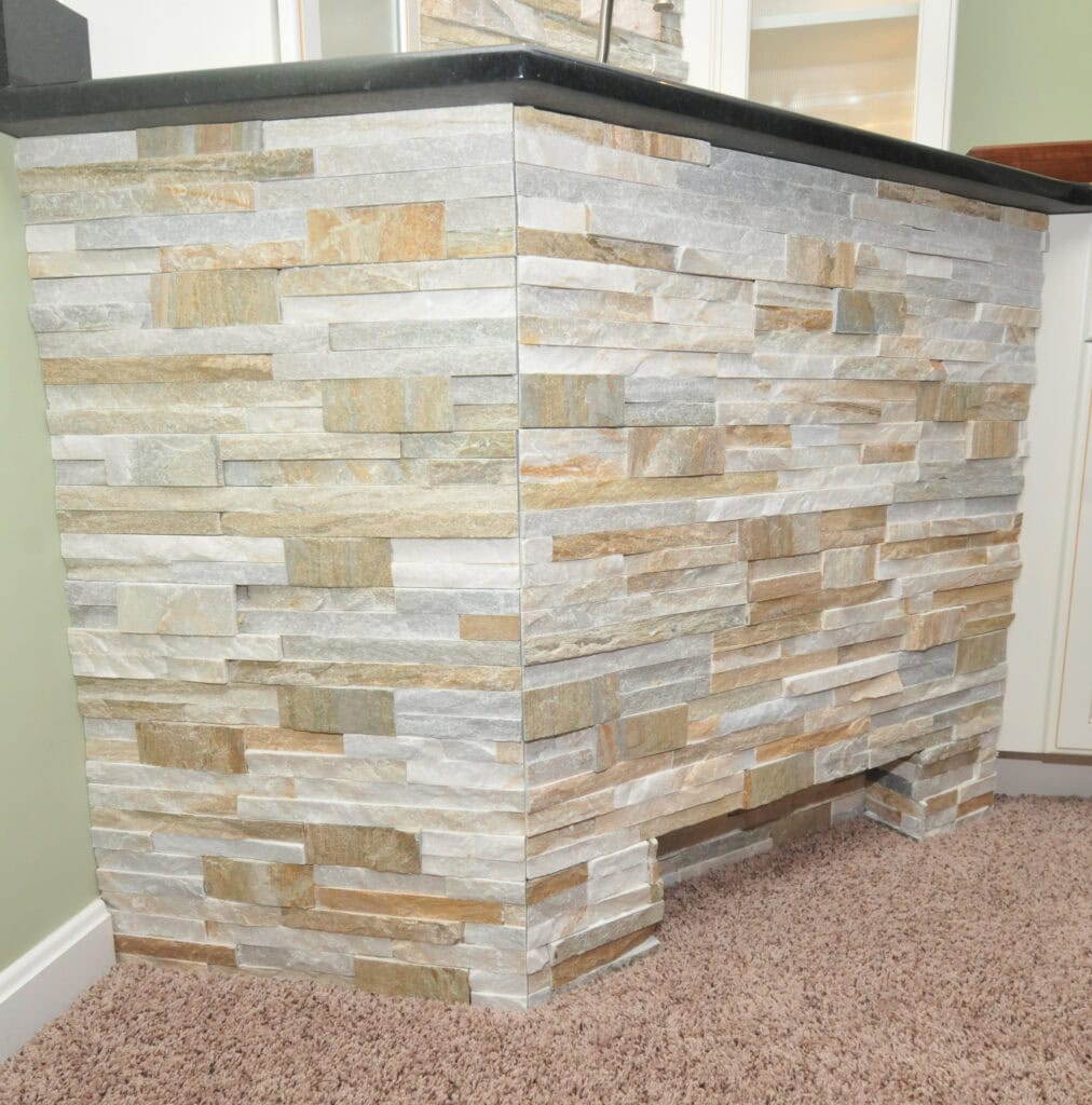 A close-up view of a bar or counter with a stacked stone veneer in shades of beige, gray, and tan, set against a carpeted floor and light green wall.