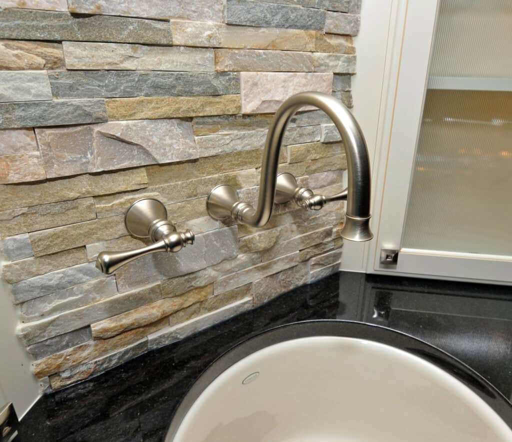 A wall-mounted faucet with two handles is installed above a round vessel sink on a black countertop, set against a decorative stone tile backsplash.