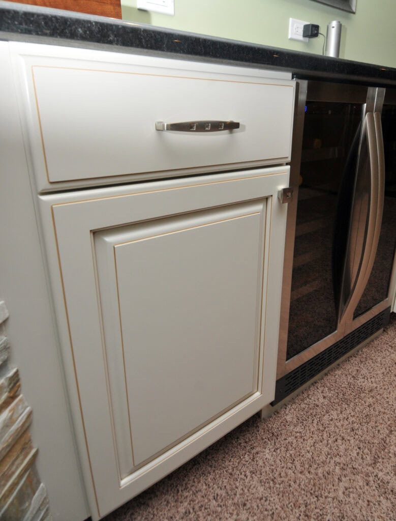 A white cabinet with a drawer and a door, featuring silver handles, is next to a stainless steel kitchen appliance under a black countertop. The cabinet is set against a carpeted floor.
