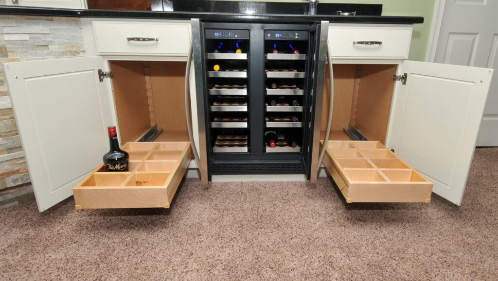 Two open cabinets with sliding wooden drawers and compartment organizers flank a dual wine cooler stocked with bottles. One drawer holds a bottle of wine, and the floor is covered in beige carpet.