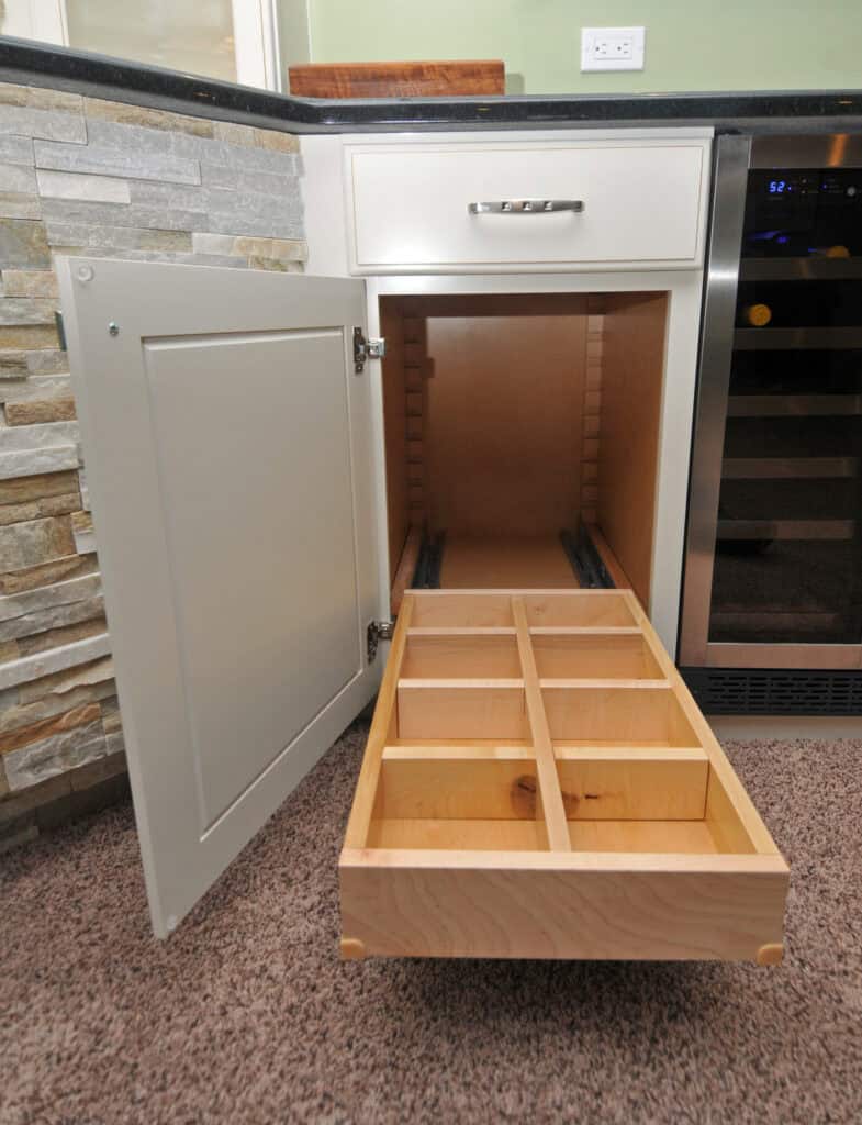 A kitchen cabinet with the door open reveals a pull-out wooden organizer with multiple square compartments, designed for storage. The cabinet is near a stone backsplash and a wine cooler.