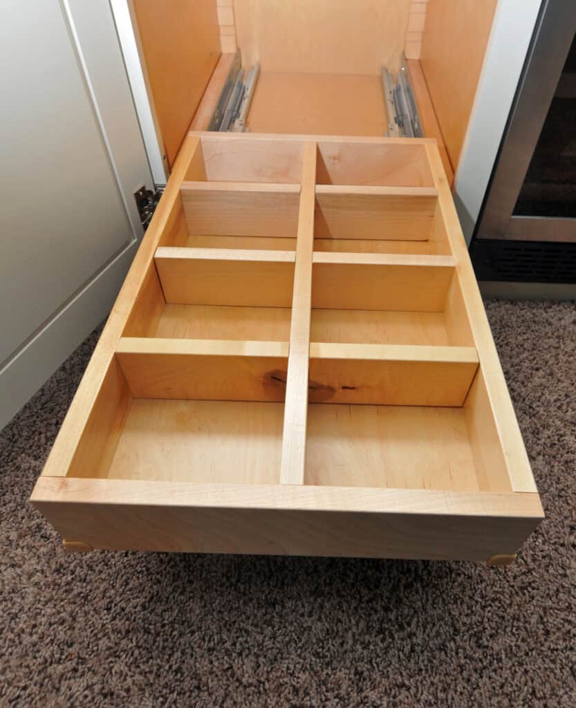 An empty wooden kitchen drawer with six rectangular compartments, designed for organizing utensils, opened above a carpeted floor.