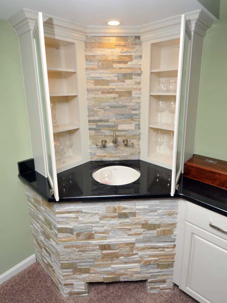 A home wet bar with a round sink, stacked stone backsplash, open corner shelves displaying glassware, black countertop, and recessed lighting, set against light green walls and carpeted flooring.