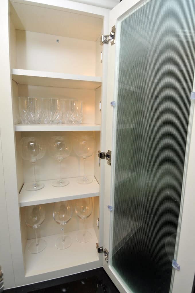 An open white cabinet with three shelves; six short crystal glasses are on the top shelf, and six large wine glasses are evenly spaced on the middle and bottom shelves. The door has a frosted glass panel.