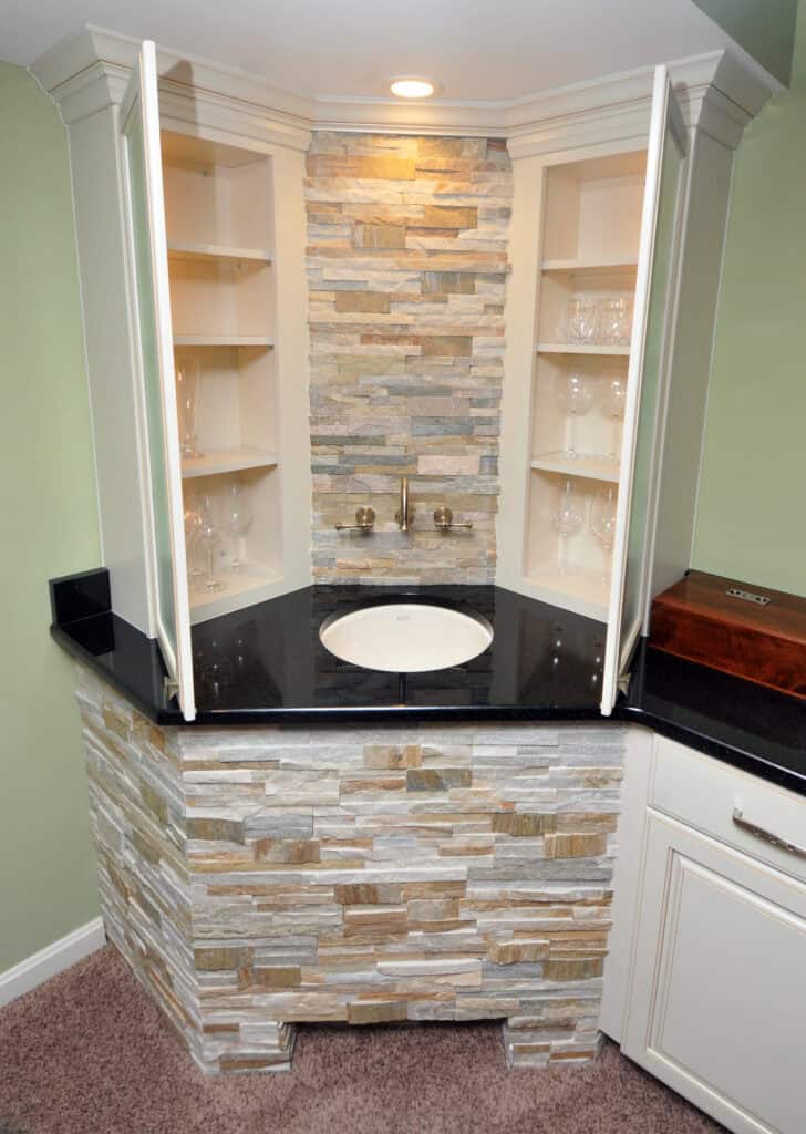 A small bar sink with a black countertop, surrounded by stone tiles, features open cabinet doors revealing glass shelves stocked with glassware. A wall-mounted faucet is set against a stone backsplash.