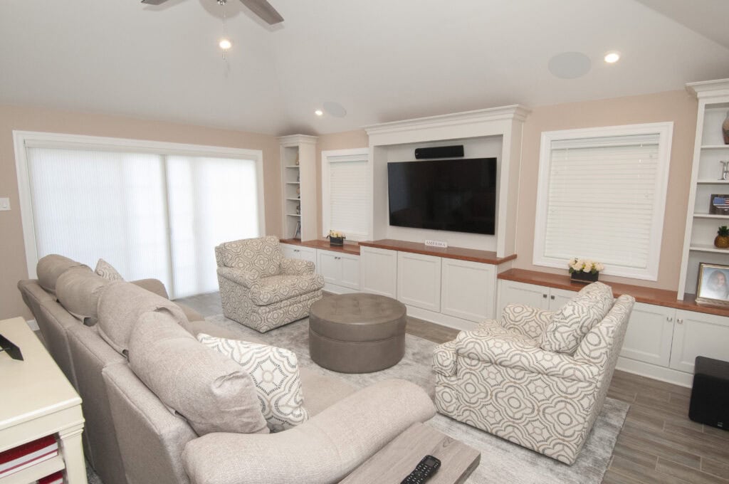 A bright living room with a large beige sectional sofa, patterned armchairs, a round ottoman coffee table, built-in white cabinets, a wall-mounted TV, and windows with white blinds.