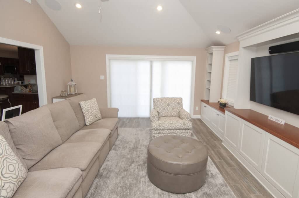A cozy living room with a beige sofa, patterned armchair, round ottoman, light gray rug, built-in cabinets with a TV, and large windows with sheer blinds letting in natural light.