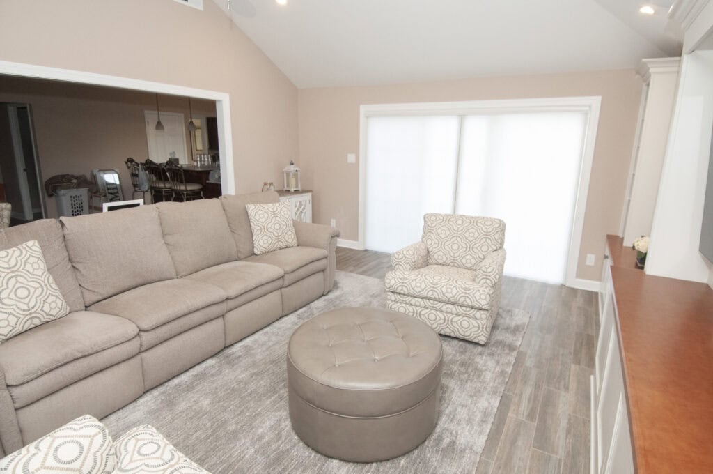 A bright living room with a beige sofa, patterned armchair, round leather ottoman, gray area rug, and light wood floors. Large sliding doors with white blinds let in natural light. A dining area is visible in the background.