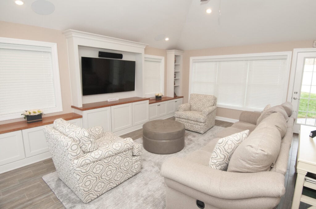A bright living room with a large TV on a built-in white entertainment center, two patterned armchairs, a beige sofa, a round ottoman, and large windows with blinds.