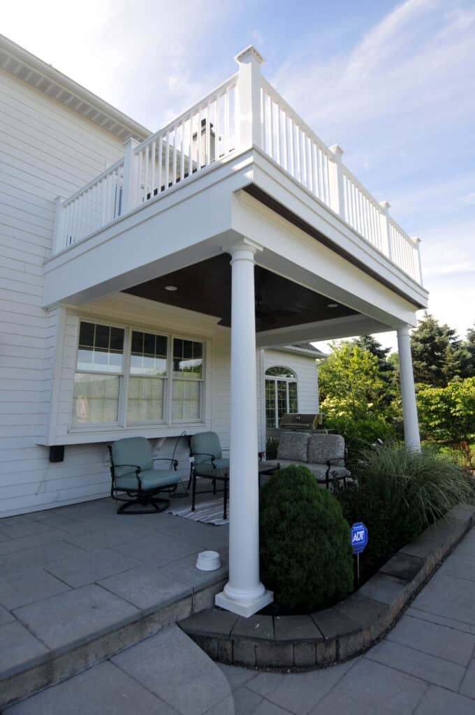 A white two-story house with a balcony supported by columns above a shaded patio. The patio has cushioned chairs, a small table, and landscaped bushes nearby under a blue sky.