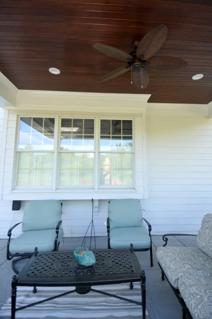 A covered patio with two light blue cushioned chairs, a patterned sofa, a black metal coffee table, a small decorative bowl, and a ceiling fan with wooden blades. Large windows are behind the seating area.