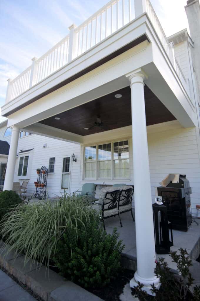 A covered patio with white columns, outdoor seating, greenery in the foreground, and an upper balcony with a white railing attached to a white house.
