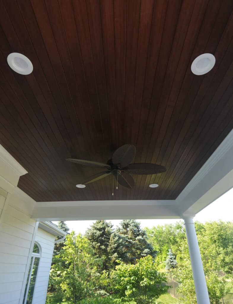 A dark wood-paneled ceiling with recessed lights and a ceiling fan featuring leaf-shaped blades, on a covered patio with white trim, overlooking a green, tree-filled yard.