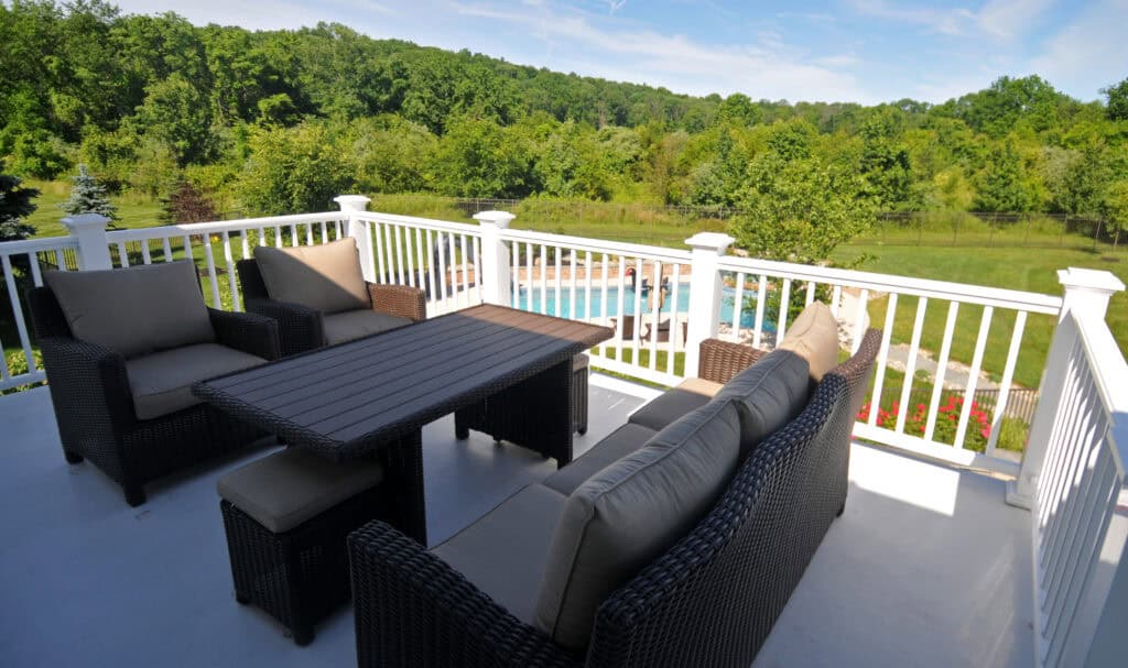 Wicker patio furniture with beige cushions sits on a deck overlooking a backyard with a swimming pool, surrounded by green trees and lawn under a blue sky.