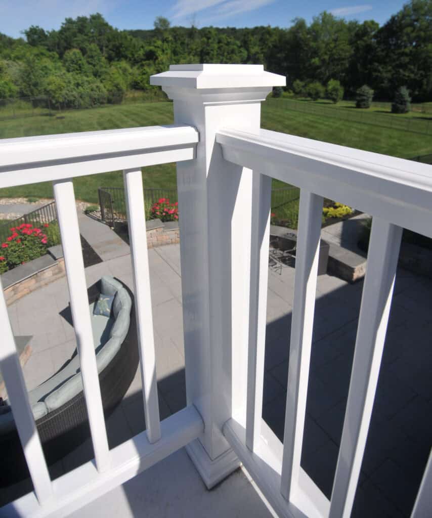 White deck railing with square post in the foreground, overlooking a patio area with outdoor furniture, garden landscaping, and a green lawn with trees in the background under a blue sky.