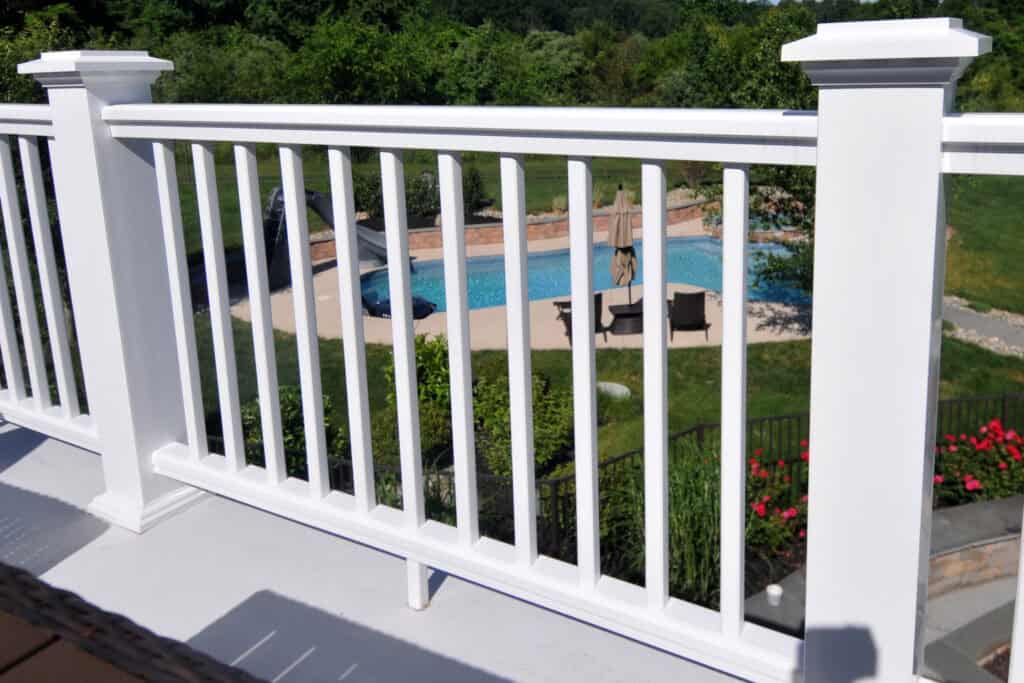 A white railing on a porch overlooks a backyard with green grass, red flowers, and a swimming pool surrounded by loungers and umbrellas. Trees fill the background.