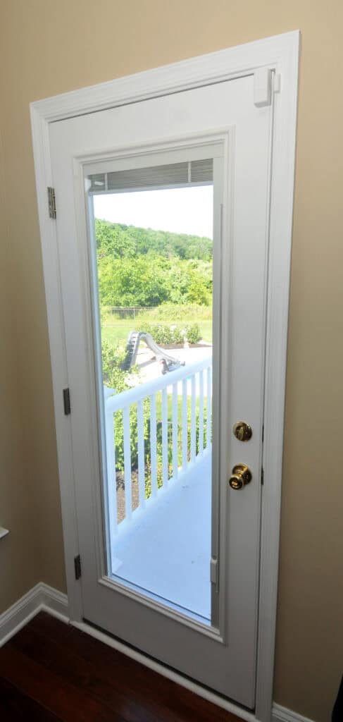 A white door with a large glass window, gold-colored doorknobs, and white trim opens to a small porch with white railing. Lush green trees and hills are visible outside under daylight.