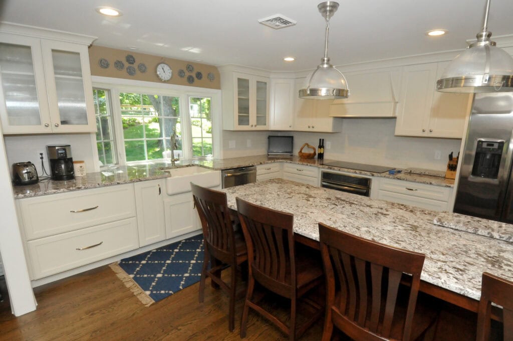 Modern kitchen with white cabinets, stainless steel appliances, granite countertops, a farmhouse sink below large windows, and four dark wooden chairs at a central island, on a hardwood floor with a blue rug.