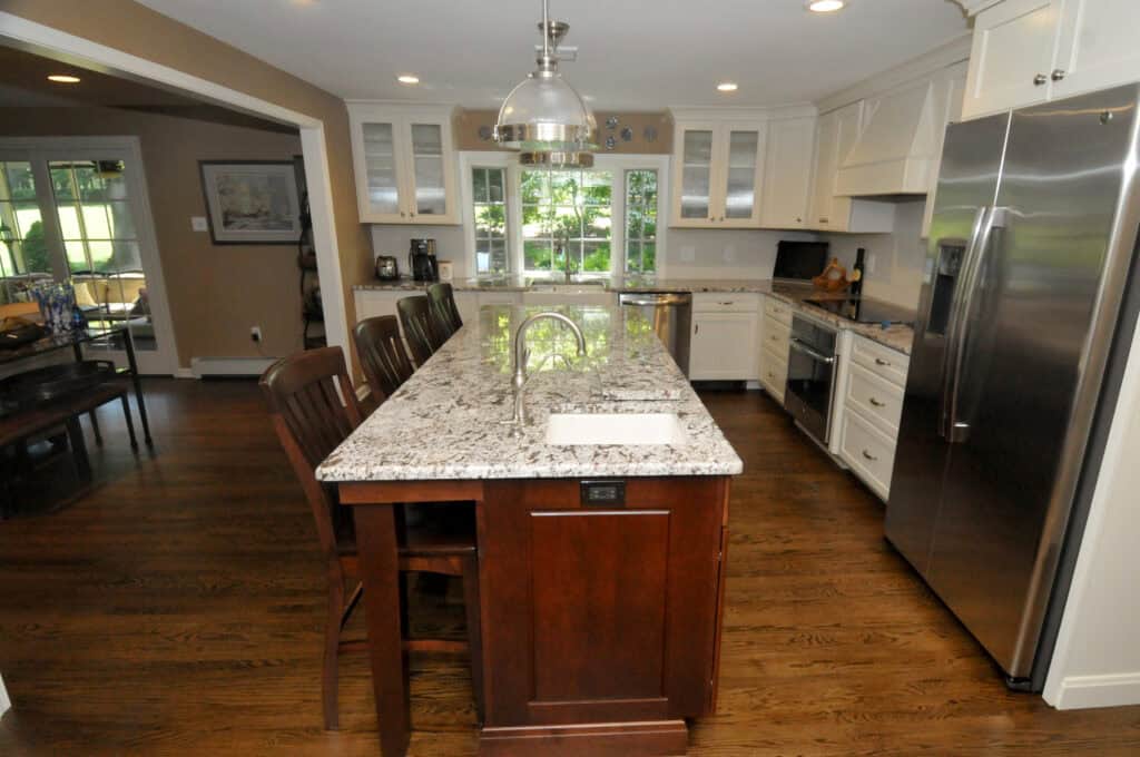 Modern kitchen with a granite island countertop, four wooden chairs, stainless steel appliances, white cabinets, and large windows letting in natural light. Hardwood floors and pendant lighting add warmth to the space.
