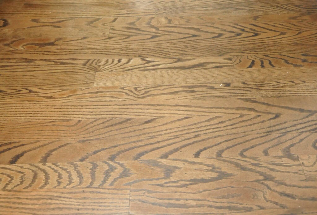 Close-up view of a wooden floor with a natural grain pattern and warm brown tones. The surface appears smooth with subtle variations in color and texture.