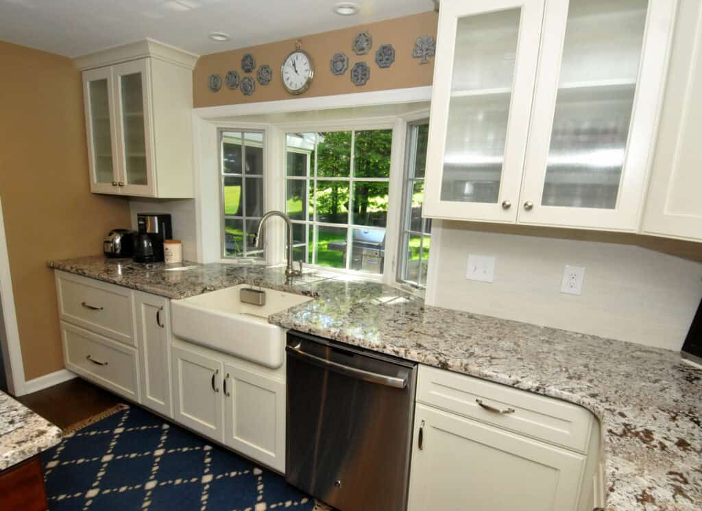 A modern kitchen with white cabinets, granite countertops, a farmhouse sink, stainless steel dishwasher, coffee maker, toaster, and a window overlooking a green backyard. Blue patterned rug on the floor.