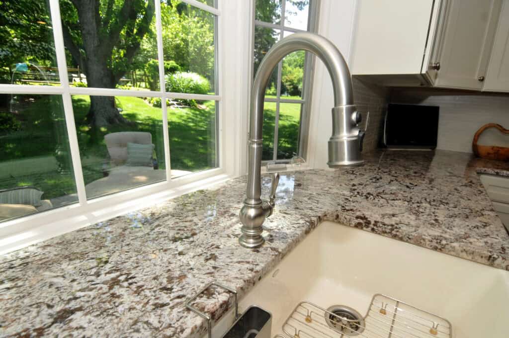 A modern kitchen sink with a stainless steel faucet sits in a granite countertop, near a large window overlooking a green backyard with trees and patio furniture.