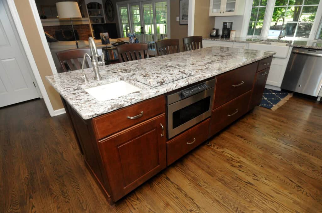 A kitchen island with a white and gray granite countertop, a built-in sink, a stovetop, a microwave, and wooden cabinets, set on a hardwood floor in a modern kitchen.