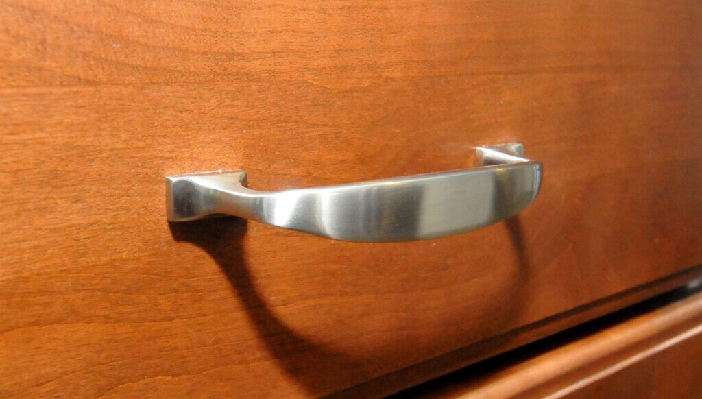 A close-up of a silver metal drawer handle mounted on a wooden drawer with a smooth, medium-brown finish. The handle casts a shadow on the drawer surface.