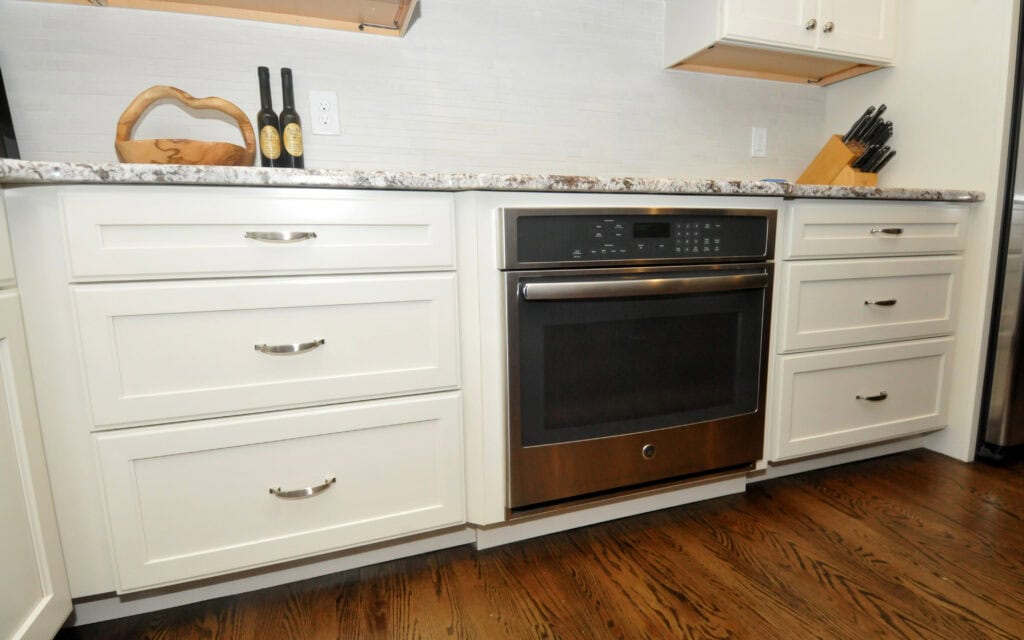 A modern kitchen with white cabinets, a built-in stainless steel oven, marble countertop, bottles of oil, a wooden cutting board, a knife block, and a dark hardwood floor.
