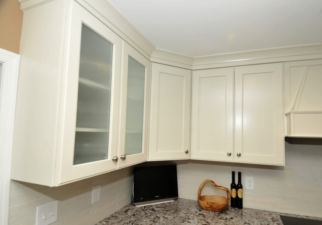 White kitchen cabinets with silver knobs are mounted above a granite countertop. On the counter are a decorative wooden bowl and two bottles, with a black appliance situated in the corner.