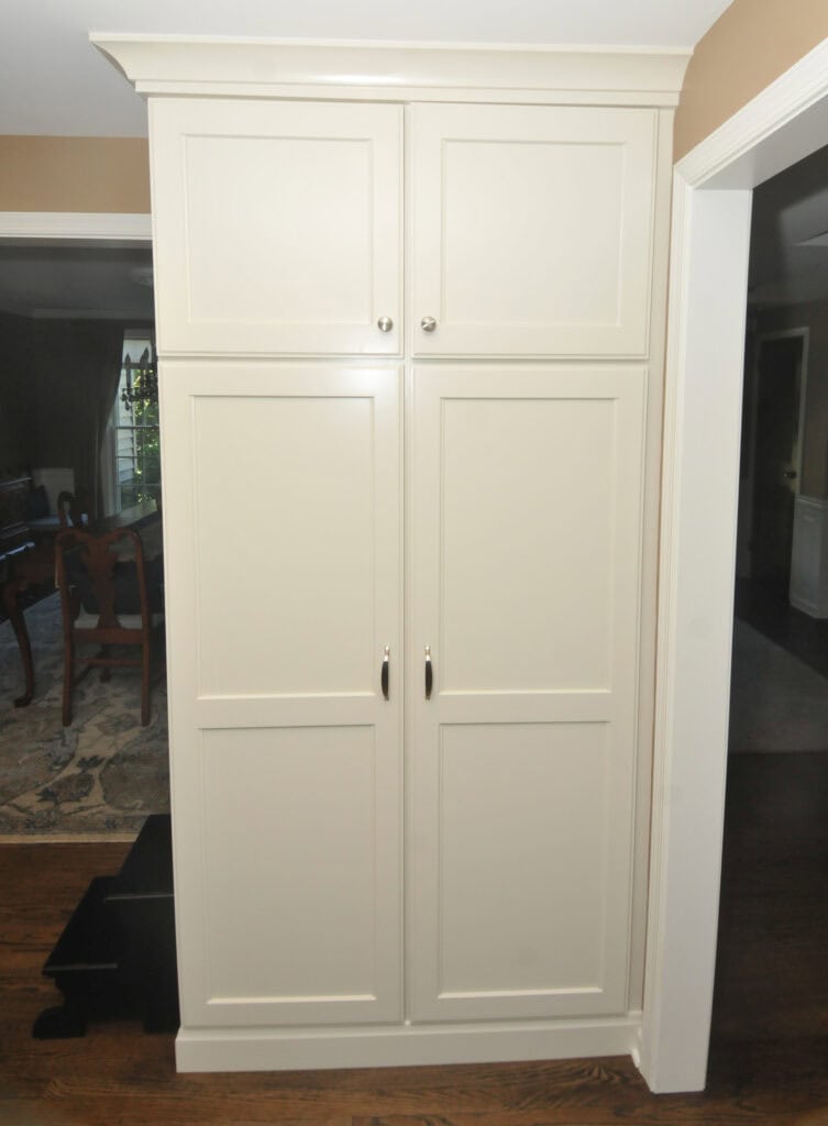 Tall, white built-in cabinet with four paneled doors and silver handles, situated between two doorways in a home with wood flooring.