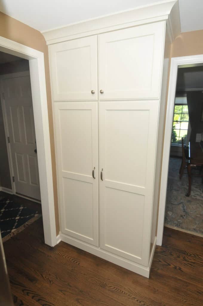 Tall, white double-door cabinet with silver handles, set against a beige wall and wood floor, positioned between two doorways in a home interior.
