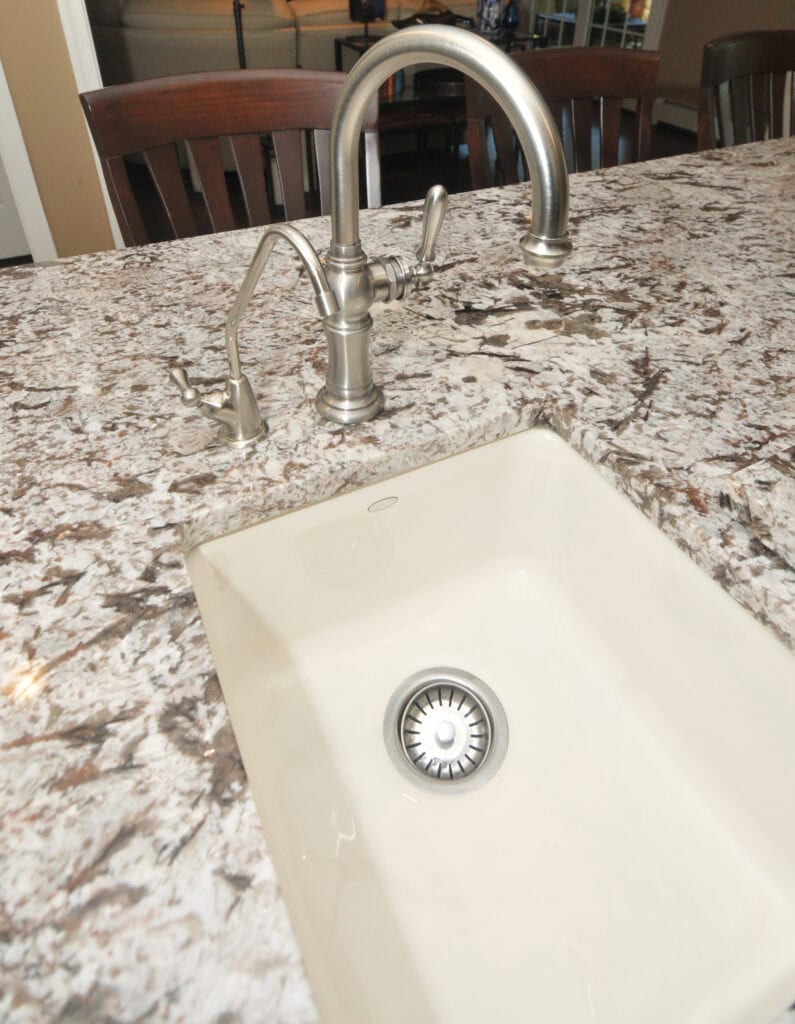A close-up of a modern kitchen sink with a brushed metal faucet and soap dispenser, set in a speckled granite countertop with dark brown wooden chairs in the background.
