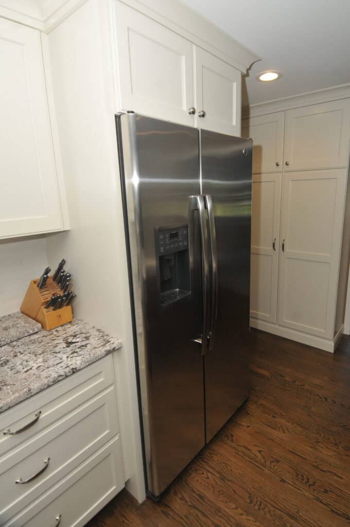 A stainless steel side-by-side refrigerator with a built-in water and ice dispenser stands next to white cabinets and granite countertops in a modern kitchen with hardwood floors.