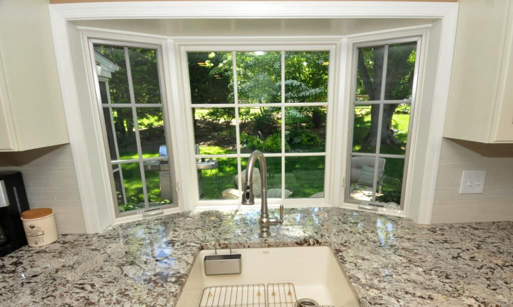 A kitchen sink with a modern faucet is set in a granite countertop, positioned in front of a large bay window overlooking a green, sunlit backyard with trees and grass.