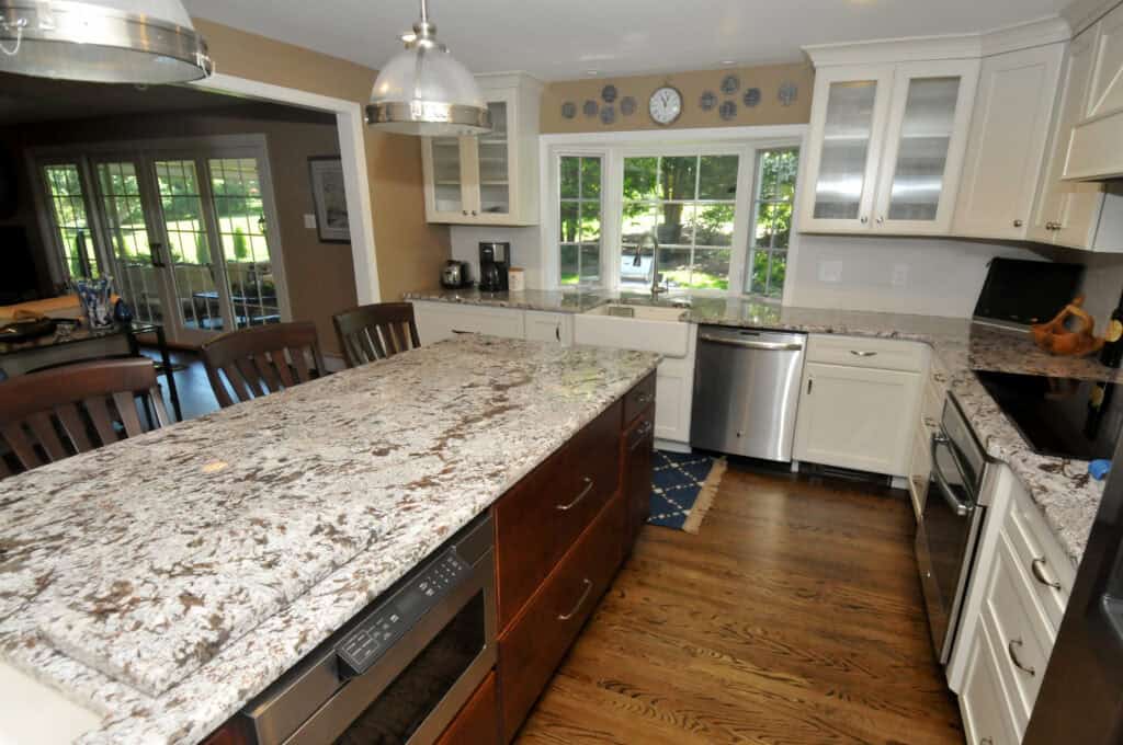 Modern kitchen with granite countertops, stainless steel appliances, white cabinets, and wooden floors. A large island with dark wood base and built-in dishwasher is in the center, with chairs along one side. Bright and spacious with windows.