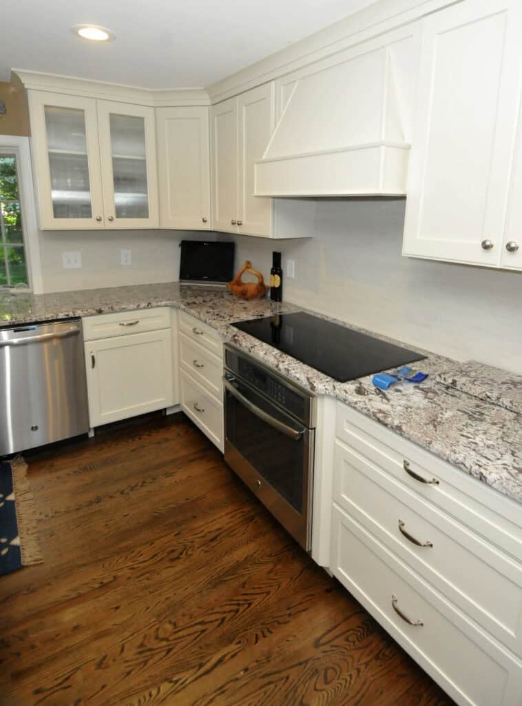 A modern kitchen with white cabinets, granite countertops, stainless steel appliances, an electric cooktop, and a built-in oven. Hardwood floors and under-cabinet lighting complete the clean, bright space.