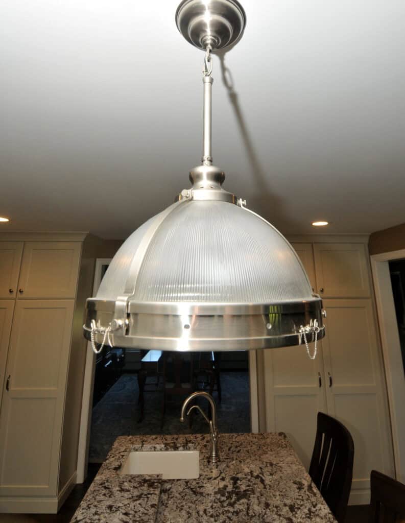 A large, industrial-style pendant light with a ribbed glass dome hangs over a granite kitchen island with a sink and faucet. White cabinetry and a dark chair are visible in the background.