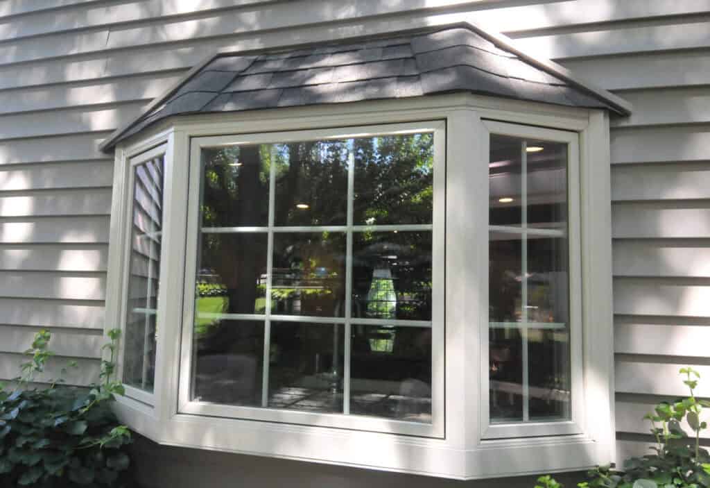 A white bay window with a shingled roof extends from the exterior of a beige house with horizontal siding. Green plants grow near the window and trees are reflected in the glass.