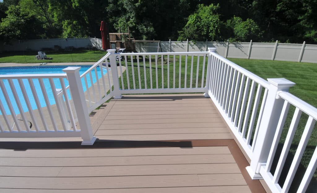 A backyard deck with white railings overlooks a green lawn and an in-ground swimming pool with a fence and trees in the background.