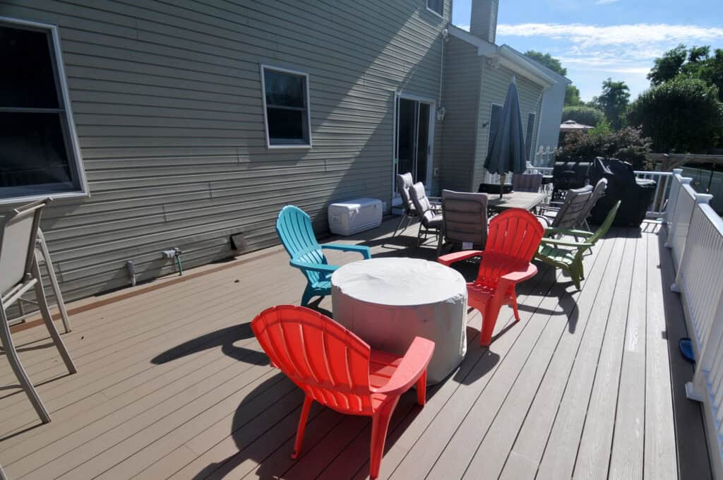 A spacious wooden deck with colorful plastic chairs around a covered table, lounge chairs, a cooler, umbrella, and a house with beige siding in the background on a sunny day.