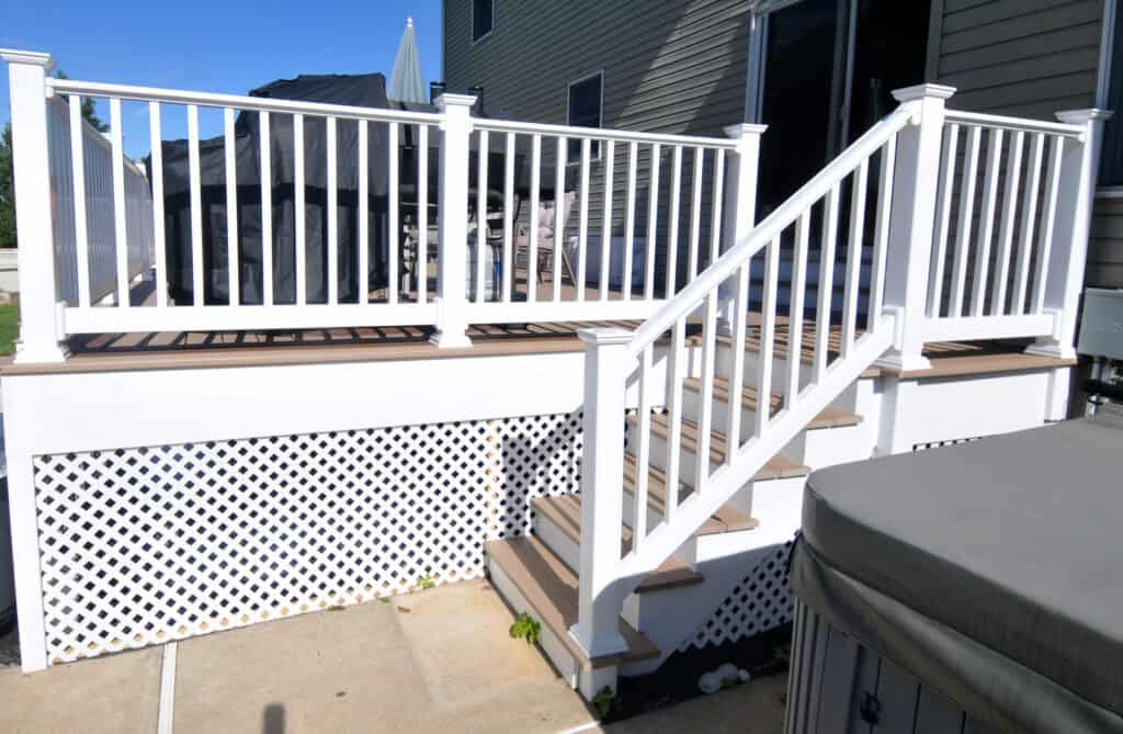 White-railed deck with lattice skirting, brown steps, and white posts, located next to a house. An outdoor grill and patio umbrella are visible on the deck. A hot tub is in the foreground.
