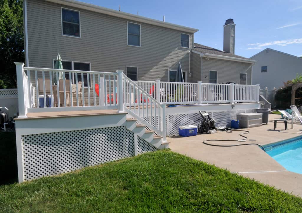 A backyard with a raised white deck, patio furniture, and steps leading down to a swimming pool. The poolside area has green grass, a hose, and a blue cooler near a tan house with large windows.