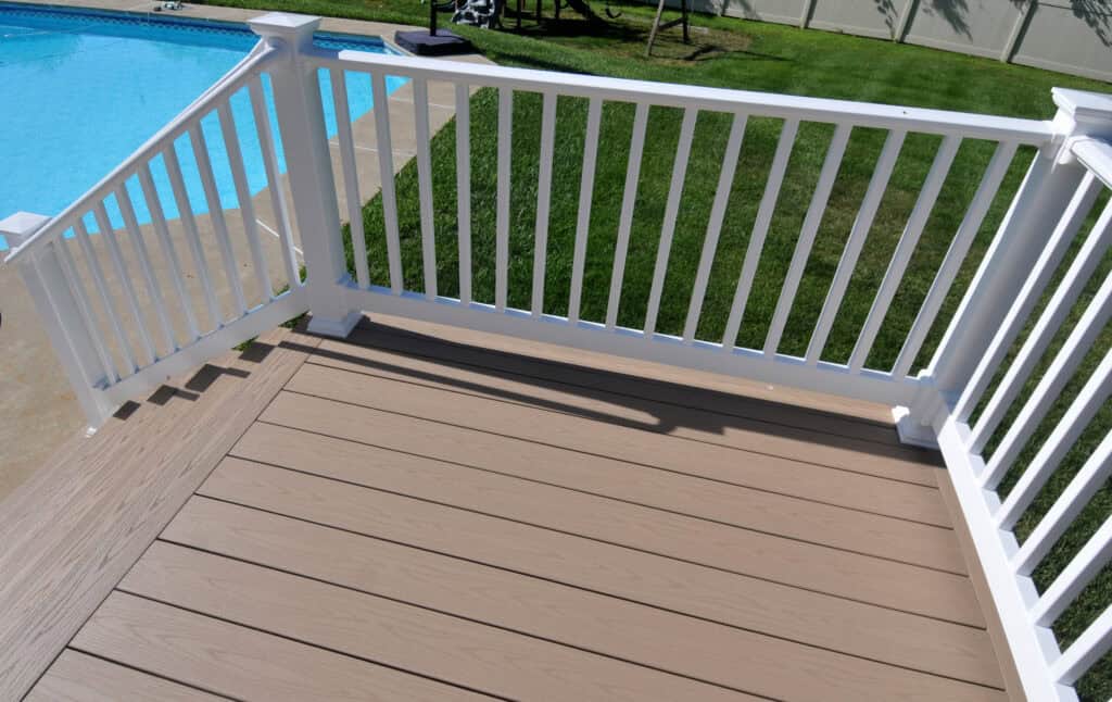 A backyard view from a deck with light brown wooden flooring and white railings, overlooking green grass and a swimming pool in the corner.
