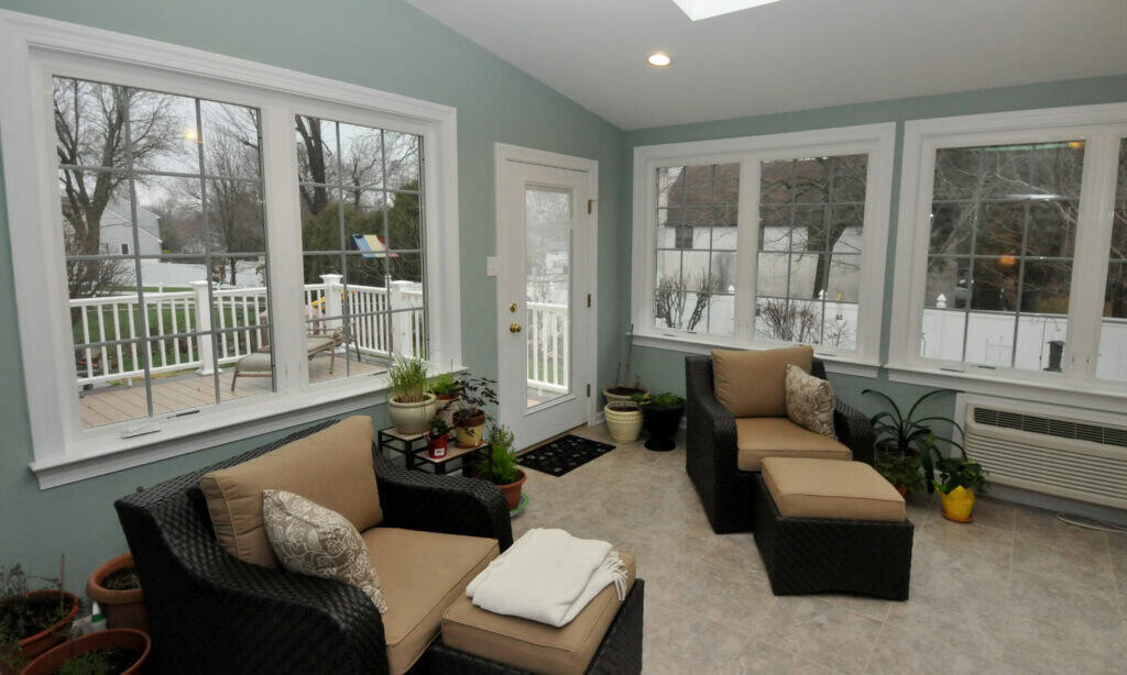 A bright sunroom with light blue walls, large windows, two wicker chairs with beige cushions, potted plants, and a door leading to a deck outside.