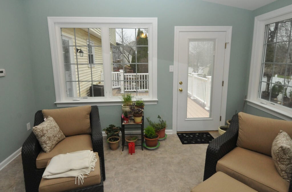 A cozy sunroom with tan cushioned chairs, a light throw, potted plants on a small stand near the window, and a glass door leading to a deck outside. Light blue walls and tiled floors complete the space.