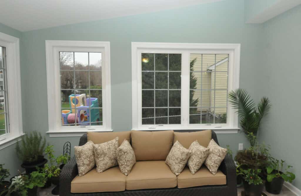A sunroom with light blue walls, a tan wicker sofa with patterned cushions, several potted plants, and large windows showing a backyard with a colorful playground outside.